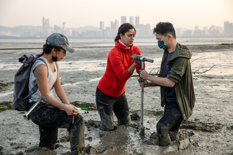 Nicole Khan and two colleagues, knee-deep in mud, collect samples from mudflats with the Hong skyline visible in distance