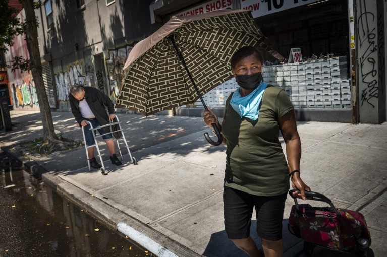 On a New York city street a woman holds an umbrella to protect her from the sun, as a man behind walks with a zimmer frame