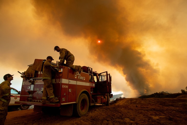 Firefighters are seen with a firetruck in the foreground while smoke rises into the sky in the background