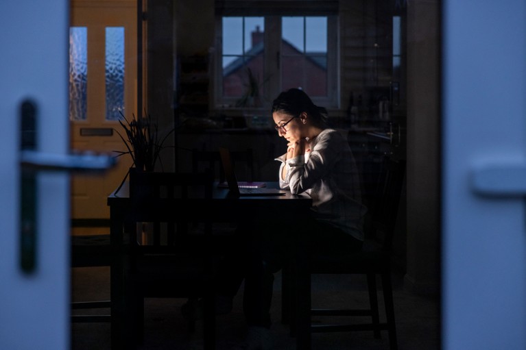 A woman is seen through a glass door working on her laptop in the dark