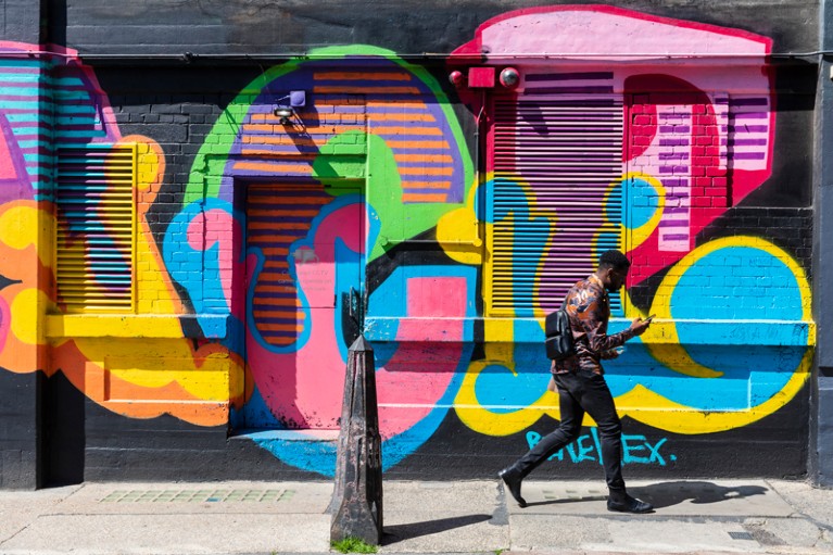 A man walks past a wall covered in colourful graffiti looking at his mobile phone