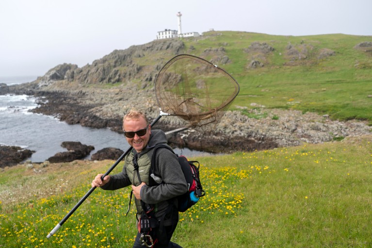 Kendrew Colhoun carrying a large net over his shoulder walks along the cliff edge of an island with a lighthouse in the distance