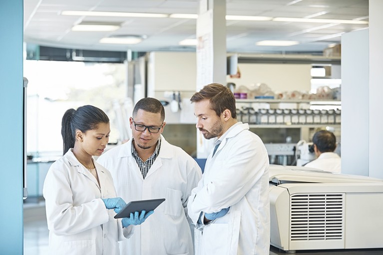 Three researchers wearing white lab coats and gloves in a lab, in discussion over a tablet.
