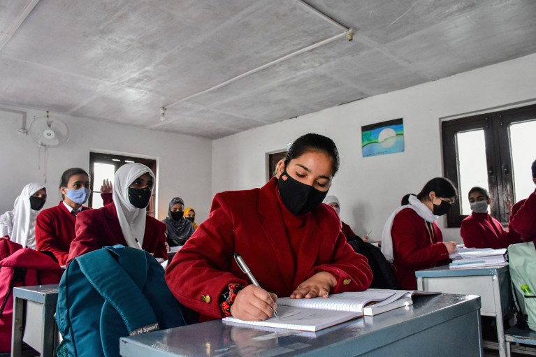 在印度的一节课上，一群十几岁的女孩戴着口罩，穿着红色运动夹克坐在课桌前