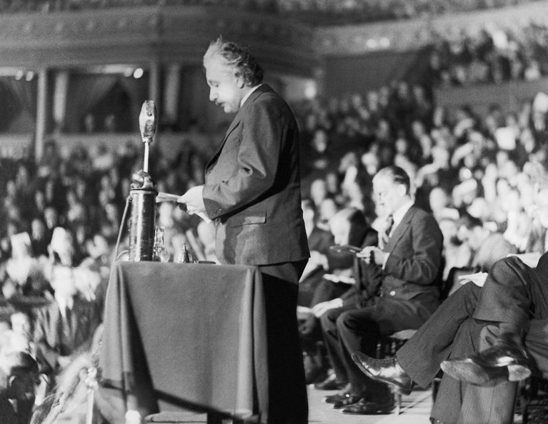 Black and white image of Albert Einstein speaking at a podium at the Royal Albert Hall, with a crowd of people behind him