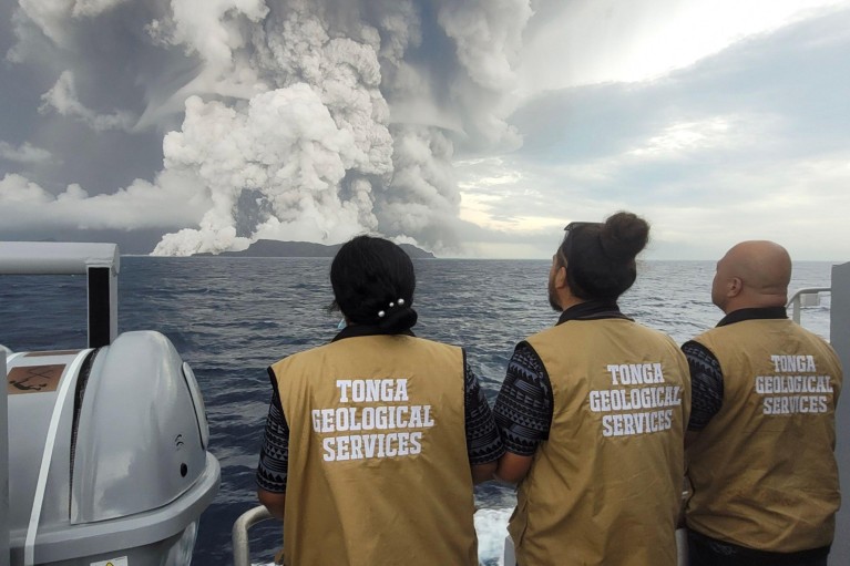 Three people wearing gold vests that read 'Tonga geological services' watch the eruption from a boat