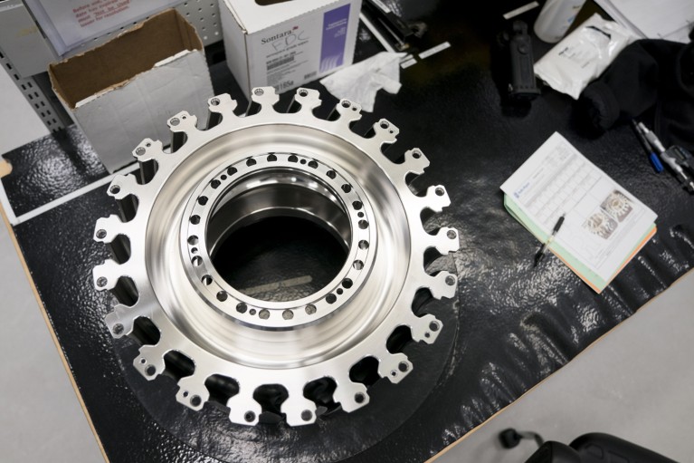 Top down view of a circular, cog-like, titanium alloy aircraft engine fan disc placed on a factory table next to some documents