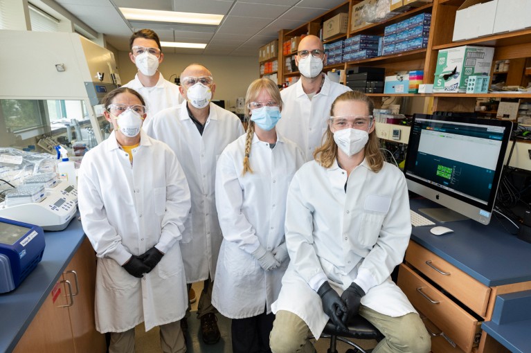 Group image of six scientists posing in the lab wearing masks, safety goggles and lab coats