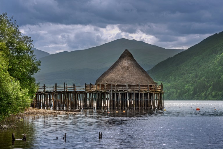 A prehistoric, thatched, cone-shaped dwelling built on a platform over a lake.