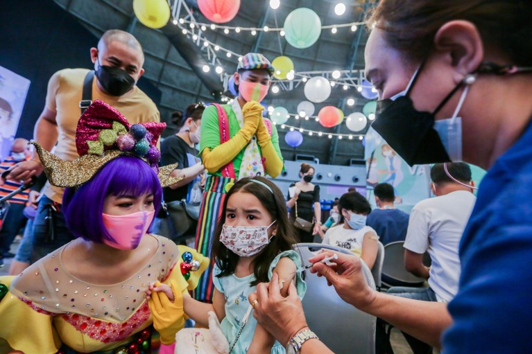 People in brightly coloured circus costumes and face masks watch as a young girl is given a COVID-19 vaccine shot into her arm