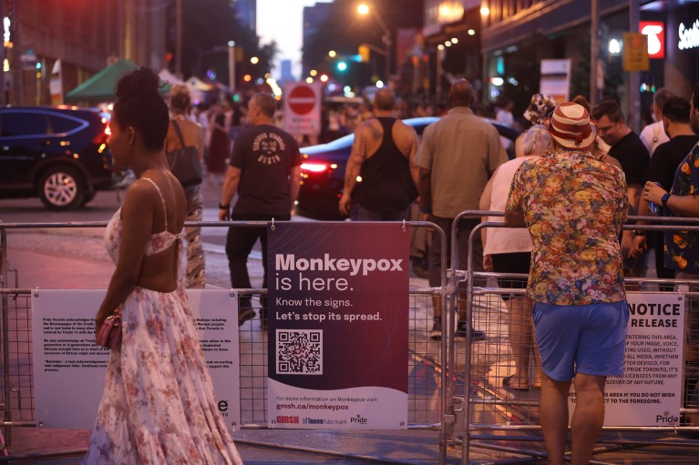 People walk past a sign reading "Monkeypox is here" attached to railings at a street event in Toronto