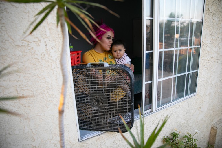 A mother holds her baby daughter by a window with fan balanced on the windowsill