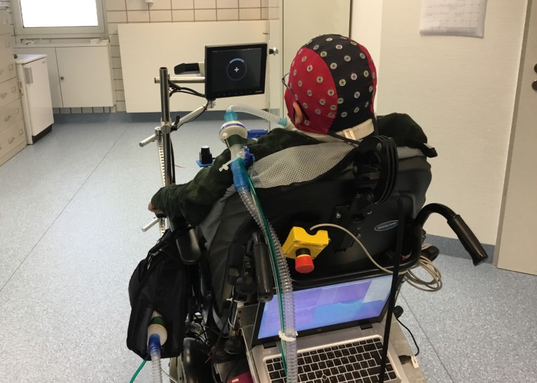 Back view of a person wearing an electrode cap, using a wheelchair attached to a computer.