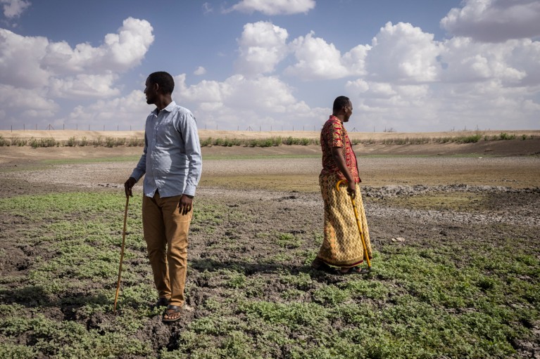 Two men holding walking sticks stand on the mud of a dried up reservoir in Kenya