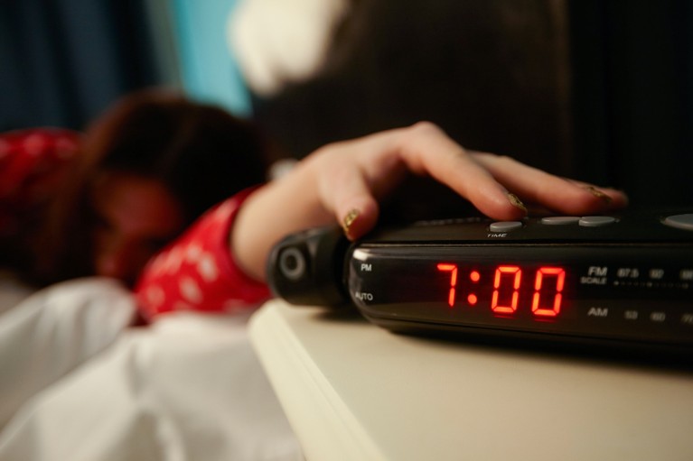 Shallow focus image of a woman reaching out of bed to turn off her alarm clock, reading 7:00