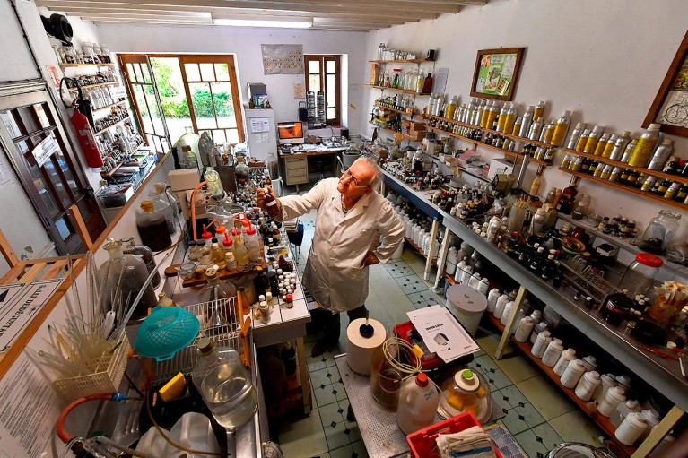 Michael Moisseeff wearing spectacles and a lab coat inspects a bottle in a wide angle view of his home laboratory