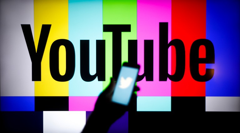 The Twitter logo is seen on a handheld mobile device with the YouTube logo displayed on a screen in the background.