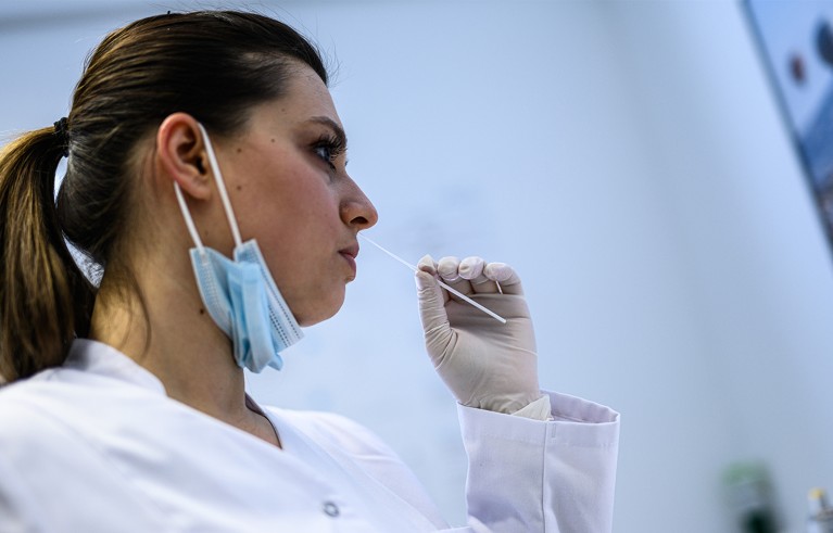 A woman in a lab coat and gloves swabs her own nose.
