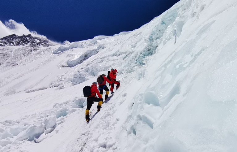 Three people in red coats with backpacks climbing a slope of thick snow.