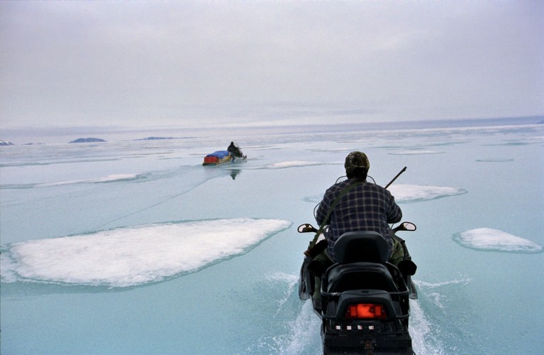 Inuits are seen on snow mobiles driving across ice that is covered in water