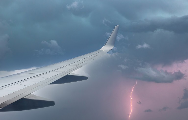 Aircraft wing slicing through a sky lit up by lightning from a thunderstorm.