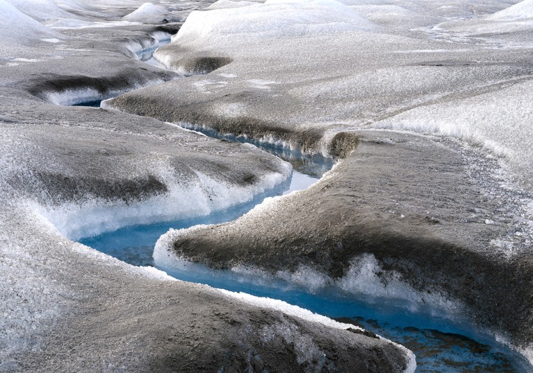 A blue river carves a channel through ice covered in brown mud.