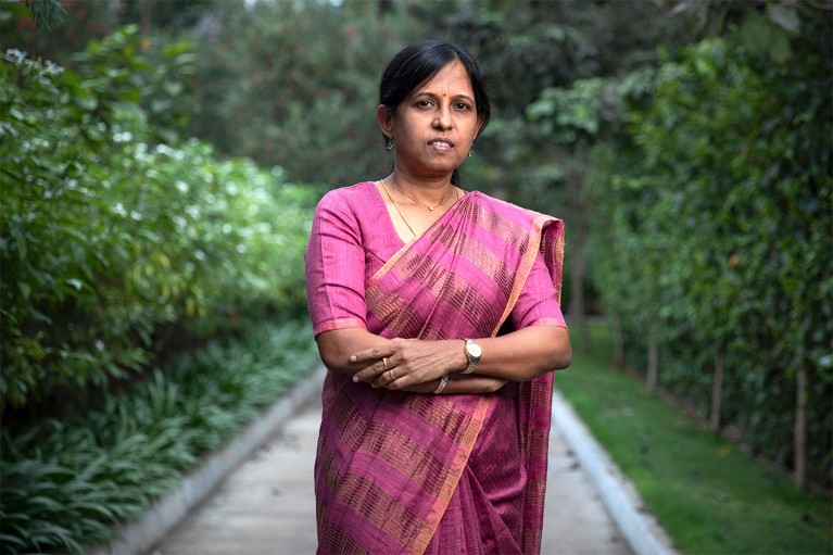 Portrait of Kalpana Kalahasti outside in a garden, her arms crossed, wearing a pink and orange sari