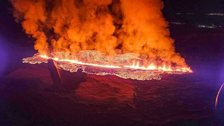 Night view of white-hot lava spewing through a fissure and flowing over the ground, emitting smoke.