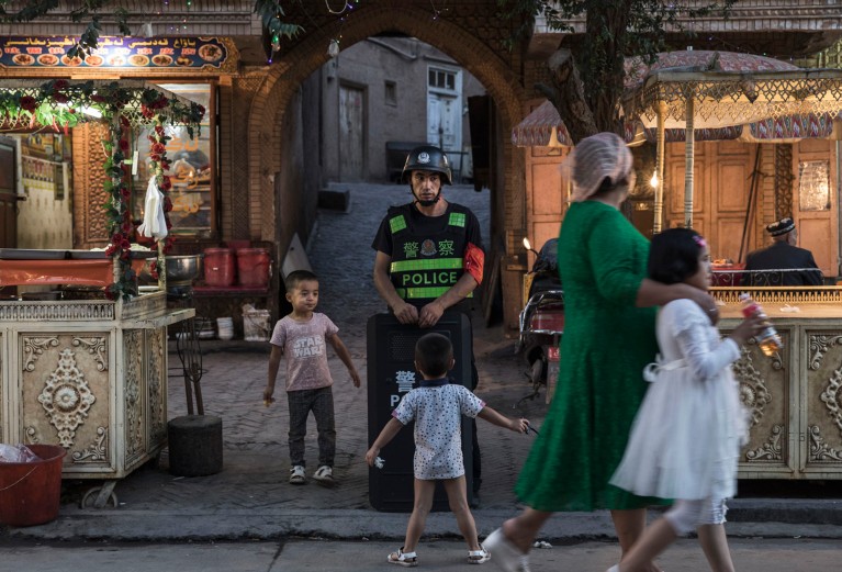 Local people walk past a policeman wearing a helmet and holding a shield in a street in Kashgar in Xinjiang province, China