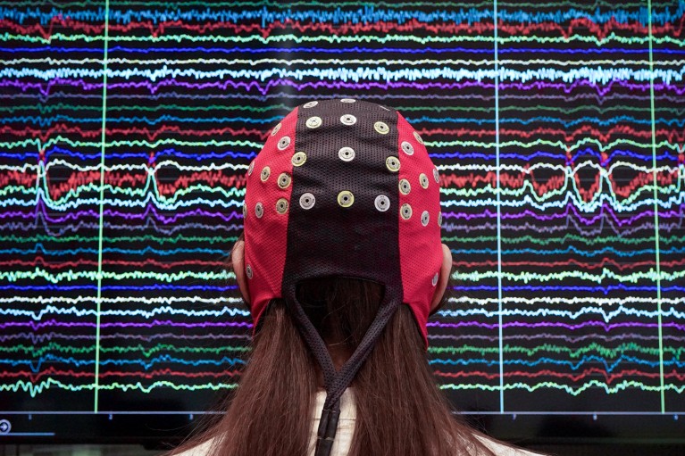 Rear-view of a woman wearing an EEG electrode cap looking at a screen covered in different coloured brain signals