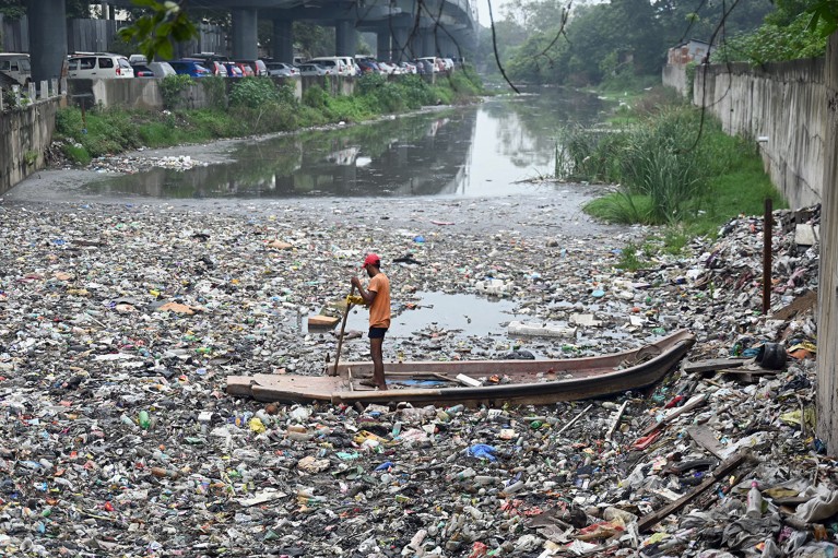 A person stands in a boat, paddling through plastic waste and litter.