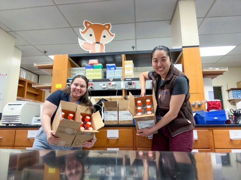 Jessica Tsai and Marissa Coppola pose for a photo in their lab while holding opened boxes of media bottles