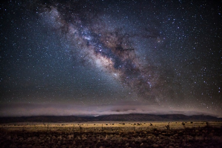A blue and purple Milky Way in the sky over a landscape of cloud-covered hills and plains in New Mexico, USA