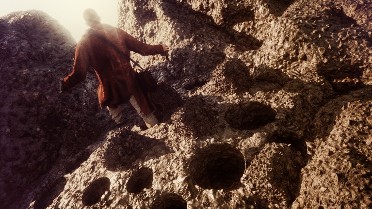 A figure wearing a roughly made tanned leather coat and with a bag on a strap over his shoulder walks through a hole-pocked rocky landscape