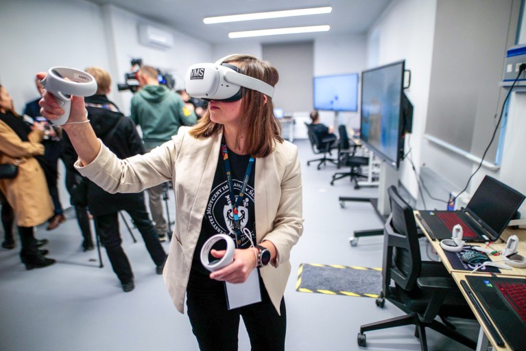 A women wearing a VR headset over her eyes and holding two controllers in a university classroom with several other people, computers and large monitors.