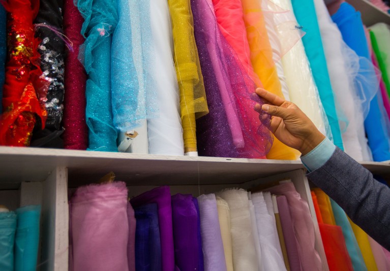 Shelves full of rolls of colourful fabrics, a man’s hand holds a purple glittery tulle.