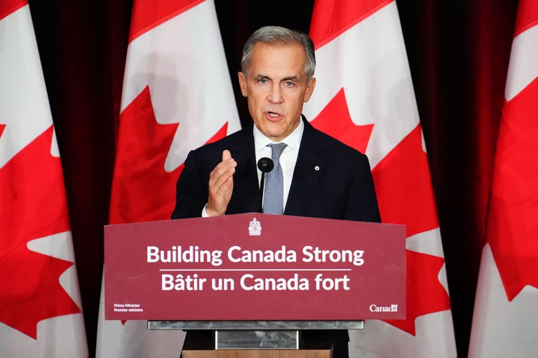 Prime Minister Mark Carney stands at a podium which reads “Building Canada Strong” behind him there is a row of red and white Canadian Flags.