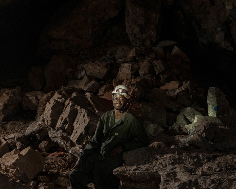 Tebogo Makhubela wearing overalls and a hard hat with a torch sitting in a dark limestone cave surrounded by large rocks