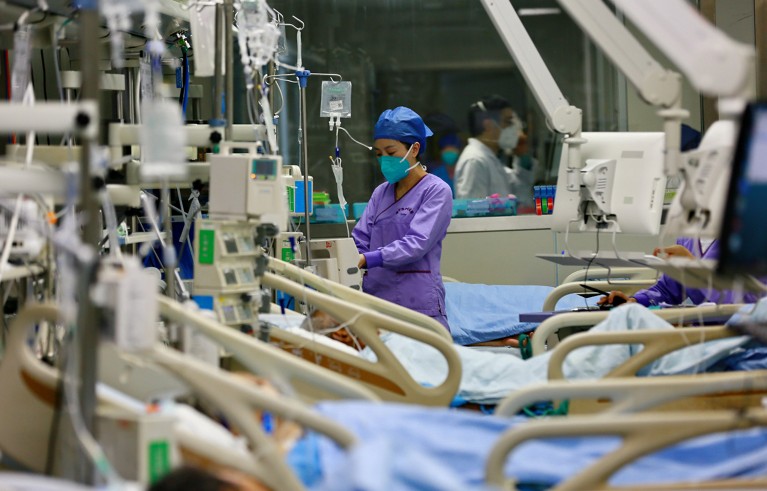 A nurse in purple scrubs and a face mask standing between ward beds.