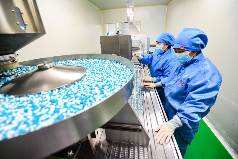 Two employees in PPE work on a production line machinery sorting medical capsules.
