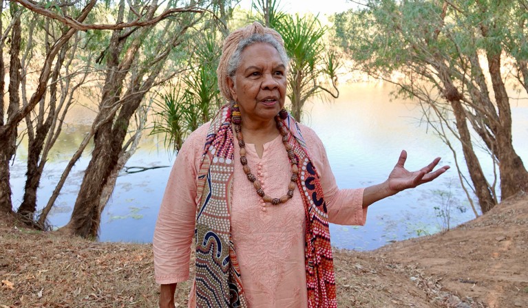 Anna Poelina stands in front of the Martuwarra Fitzroy river in the Kimberley region of Western Australia.The still water of the river behind her reflects warm golden hues. Her hand left gestures as she speaks. She wears a peach top with matching peach head wrap holding her silver hair up. A scarf with an aboriginal dot painting design hangs around her neck, alongside a wooden calved necklace and matching earings.