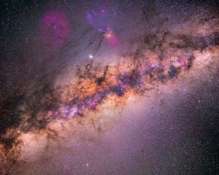 The Milky Way galaxy stretches diagonally across a star-filled sky, with dense star clusters, dark dust clouds and glowing pink and purple nebulae visible throughout