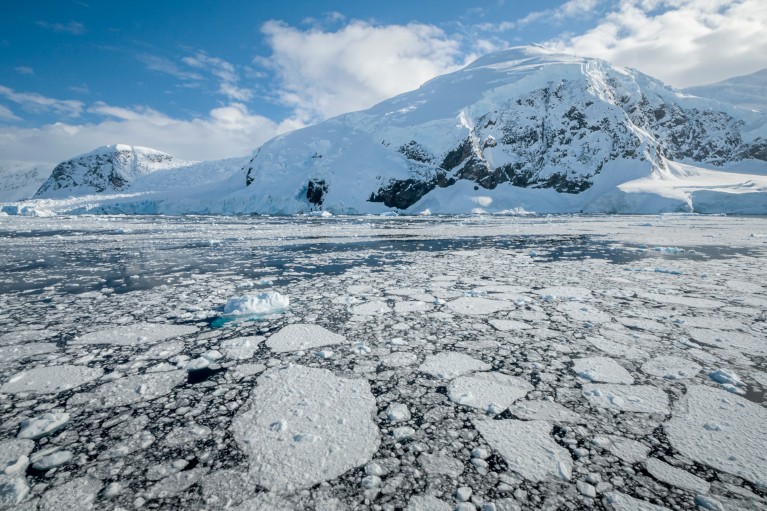 Snow-covered mountains rise above a frozen polar landscape, with fragmented sea ice floating across dark water in the foreground on a sunny day.