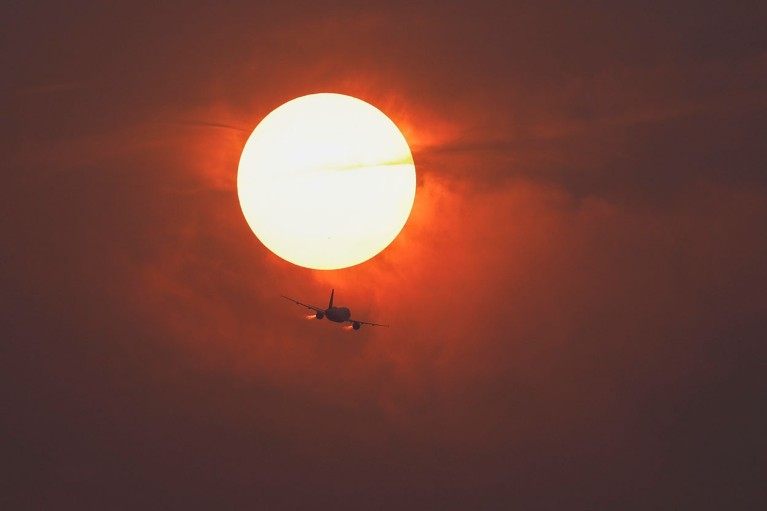 Silhouette of an aeroplane flying beneath a large bright Sun in a red-orange sky.