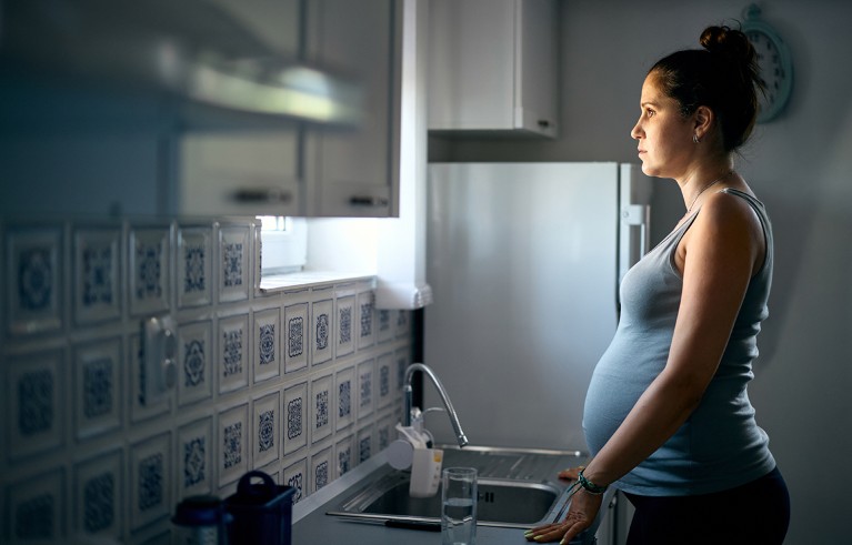 A pregnant woman standing at a kitchen sink in a softly-lit room.