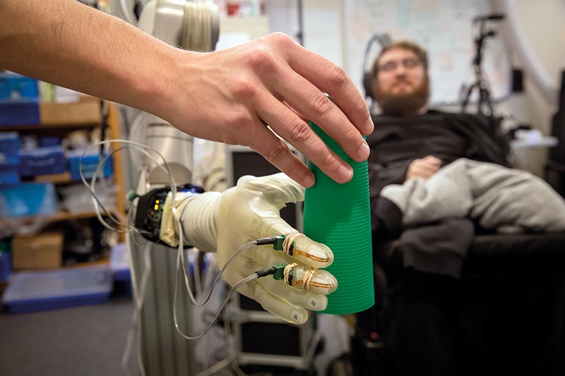 An object is placed in a robotic hand (foreground) that is connected through electrodes to Nathan Copeland (background).